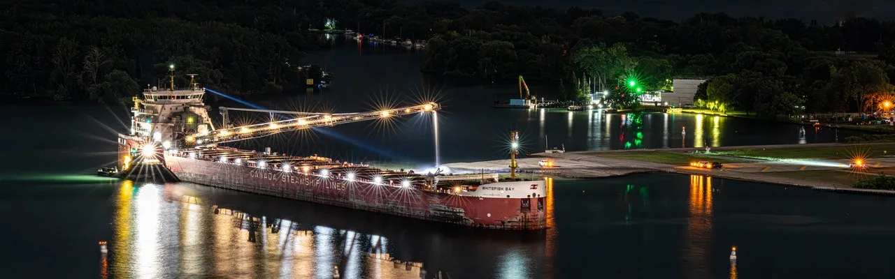 Le vraquier Whitefish Bay illuminé, accosté au quai du Port de Toronto la nuit, sa flèche de déchargement déployée au-dessus de la jetée et les lumières du havre reflétées dans l’eau.