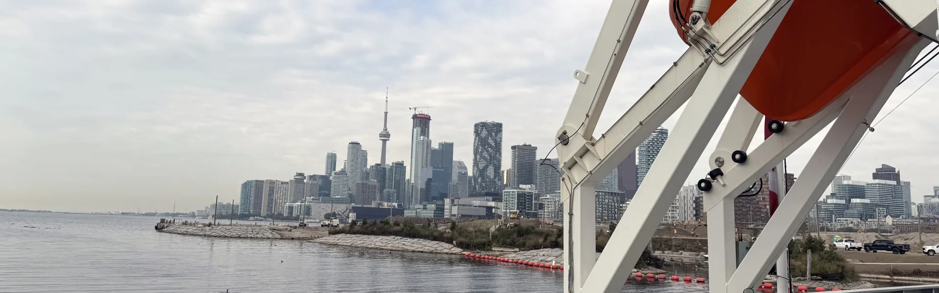 Le pont d’un navire de l’Administration portuaire de Toronto, avec des bittes d’amarrage et une embarcation de sauvetage au premier plan et la ligne d’horizon de Toronto de l’autre côté du havre.
