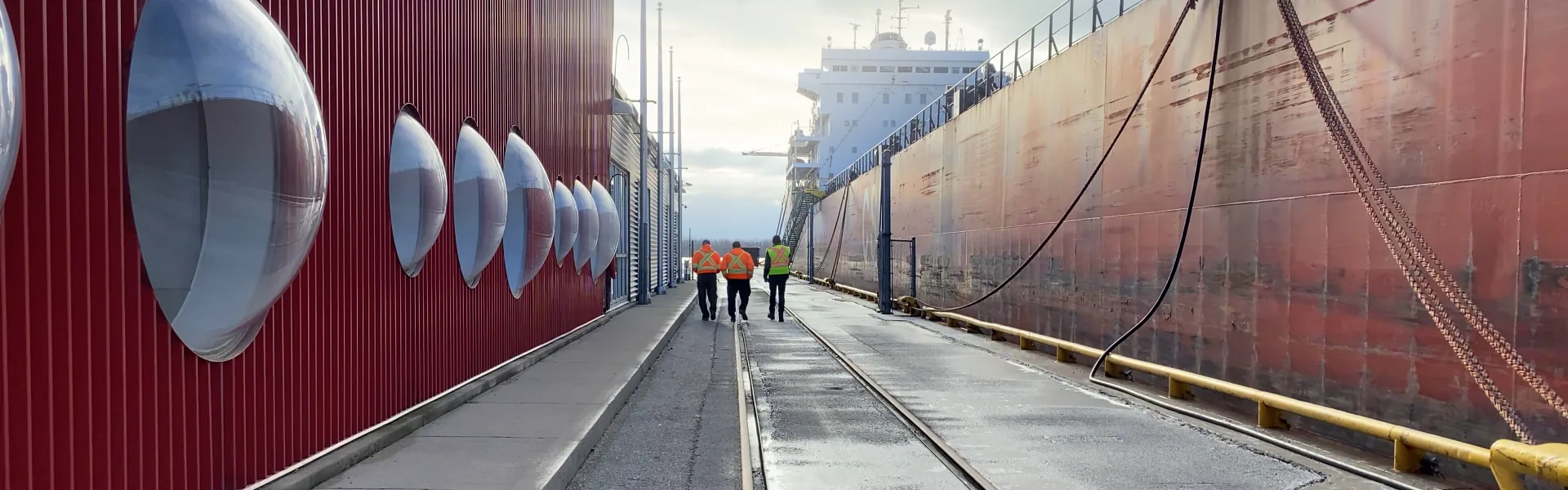 Trois employés portant des vestes de haute visibilité marchent sur le quai du Port de Toronto, le long d’un navire de charge amarré.